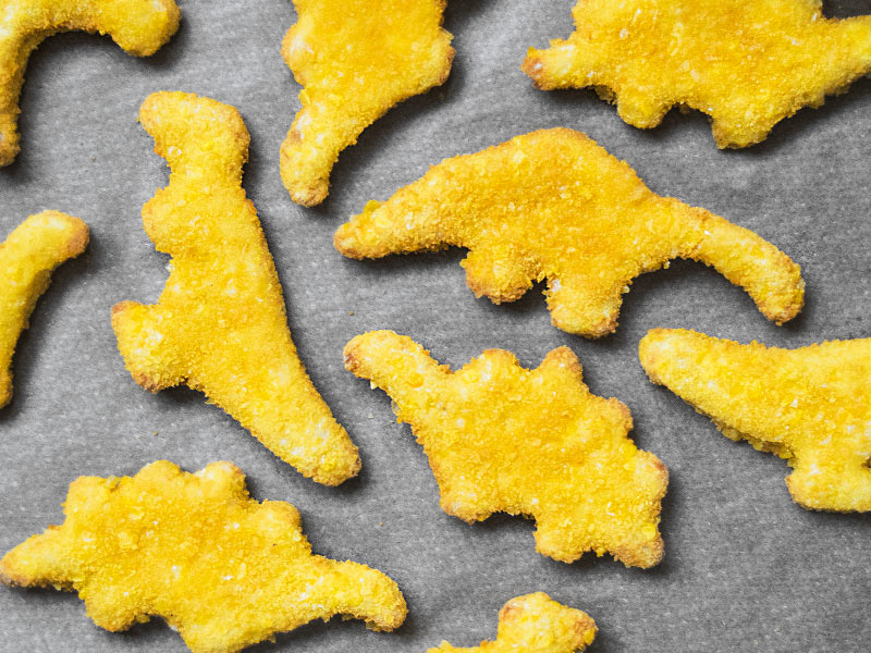 Some oven-baked chicken nuggets in the shapes of dinosaurs, laid out on a baking tray